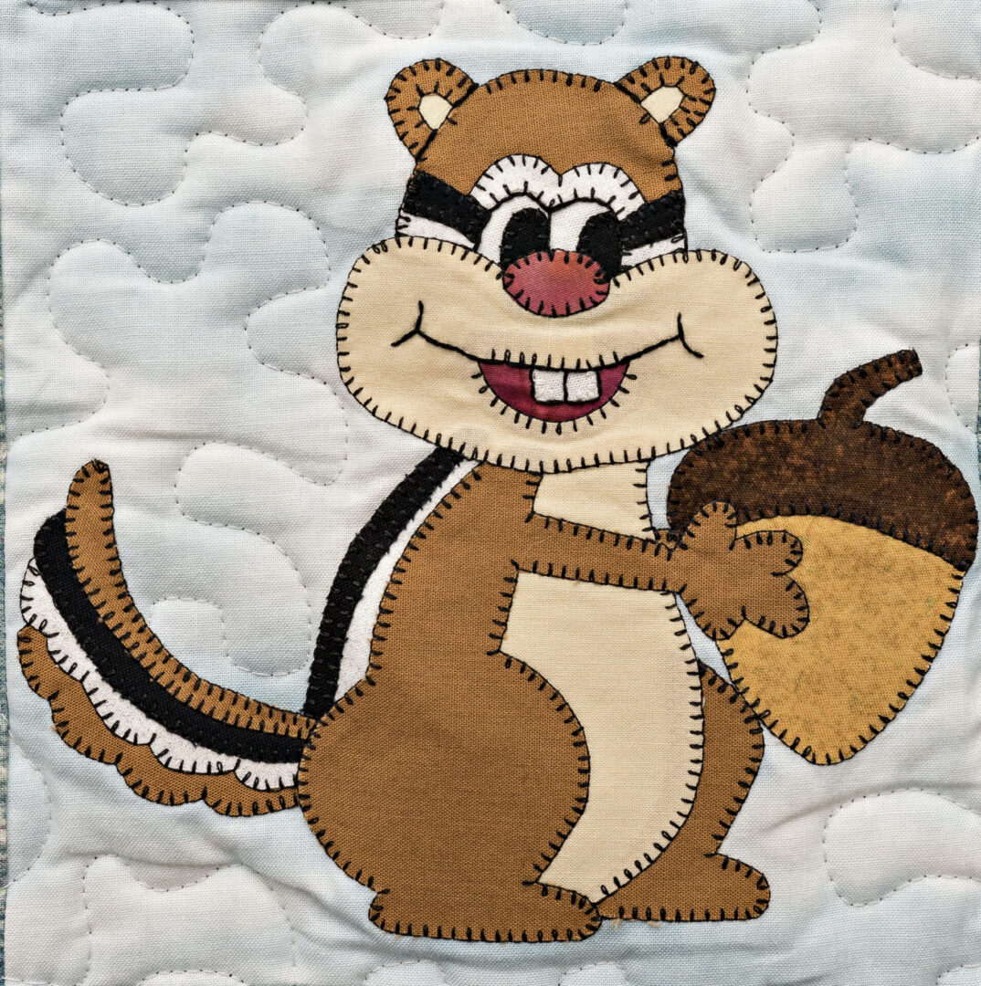 A chipmunk holding an acorn on a quilted wall hanging.