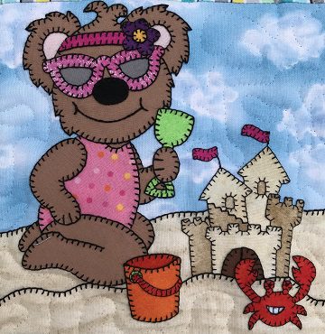 A Sand Castle Bear with sunglasses and a crab on the beach.