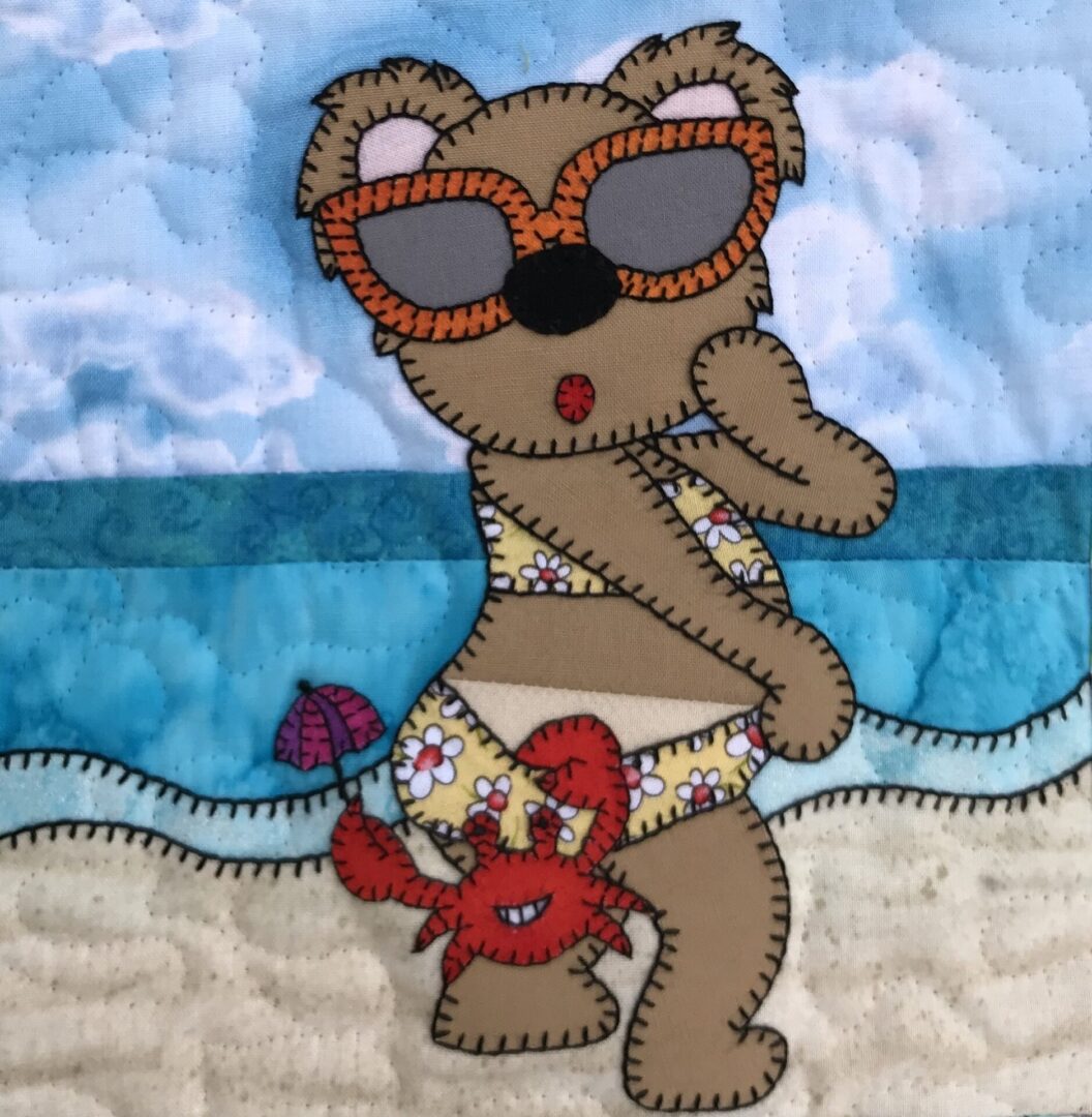 A quilt with a Suntan Bear wearing sunglasses on the beach.